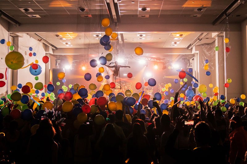 balloons floating above a crowd. circus performer in the distance suspended from the ceiling.